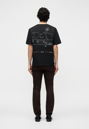 Homme portant un t-shirt noir avec un dessin au trait blanc représentant une maison, une personne, un arbre et un vélo au dos, associé à un pantalon foncé et des chaussures noires.