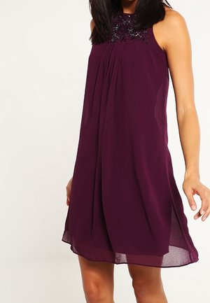 Femme portant une robe en chiffon violet foncé sans manches avec un détail décoratif au niveau du décolleté, debout devant un fond clair uni.