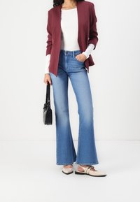 Blazer marron, chemise blanche à manches longues, jeans évasés bleu clair, chaussures beige à enfiler avec des clous, et un sac à main noir pour le contraste.
