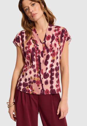 Kobieta nosząca bluzkę z krótkim rękawem w kształcie litery V z różowo-burgundowym i pomarańczowym abstrakcyjnym wzorem florystycznym, zestawioną z burgundowymi spodniami i złotymi bransoletkami.