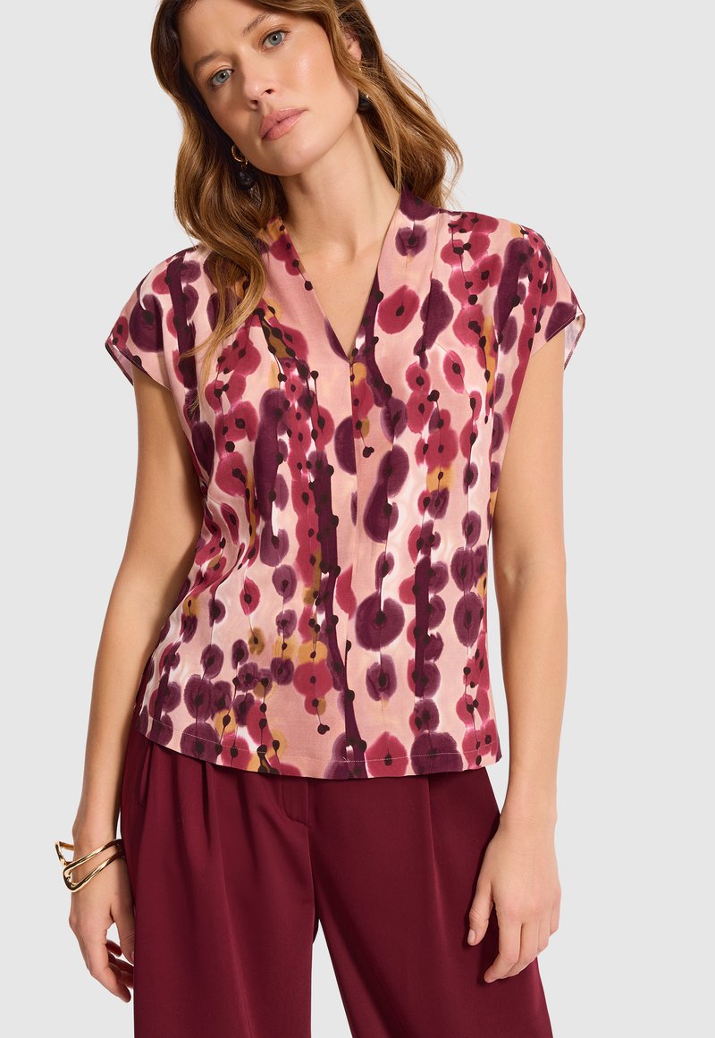 Kobieta nosząca bluzkę z krótkim rękawem w kształcie litery V z różowo-burgundowym i pomarańczowym abstrakcyjnym wzorem florystycznym, zestawioną z burgundowymi spodniami i złotymi bransoletkami.