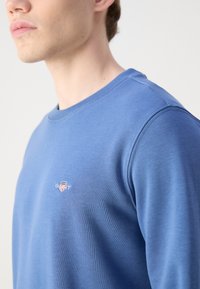 Young man wearing a light blue sweatshirt with small embroidered logo on the left chest, facing slightly left against a white background.