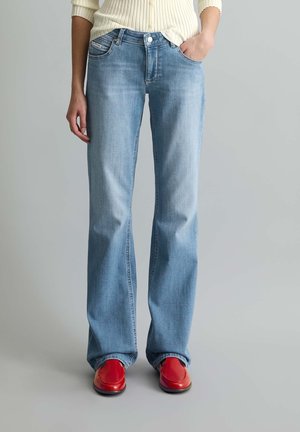 Person trägt hellblaue Jeans mit geradem Bein und rote Loafers, eine Hand in der Tasche, am Saum ist ein cremefarbener, gerippter Pullover sichtbar.