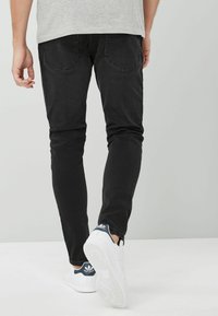 Juodieji slim-fit džinsai, pagaminti iš džinsinio audinio, su užpakalinėmis kišenėmis, subtiliais siūlės detalėmis ir glotnia tekstūra, derinami su baltais sportbačiais.