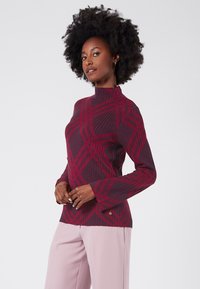 Maglione bordeaux con collo alto, texture a coste e motivo geometrico rosso. Maniche lunghe, silhouette aderente e accento del logo discreto.