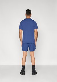 Μπλε αθλητική μπλούζα με κοντά μανίκια και στρογγυλή λαιμόκοψη, συνδυασμένη με ταιριαστό μπλε σορτς. Φοριούνται μαύρα αθλητικά παπούτσια και κάλτσες.