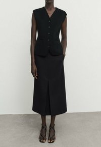 Gilet sans manches noir au design ajusté, avec cinq boutons et deux poches avant, associé à une longue jupe noire avec une fente centrale.