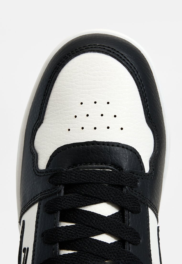 Zapatilla negra y blanca con puntera perforada y cordones negros, mostrando cuero texturizado y costuras detalladas sobre un fondo blanco.