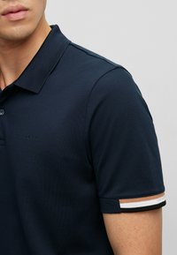 Tamsiai mėlynas polo marškinėliai su tekstūruotu audiniu, turintys subtilų logotipą ir kontrastingus dryžuotus akcentus ant rankogalių.