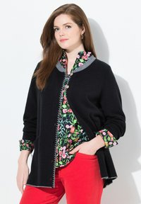 Femme aux longs cheveux roux portant des boucles d'oreilles en or, un cardigan noir, une blouse florale et un pantalon rouge, se tenant devant un fond blanc.