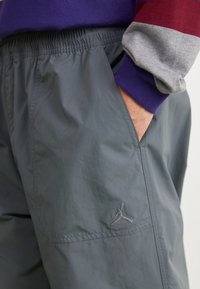 Grå joggingbyxor med elastisk midja, sidfickor och liten broderad logotyp. Materialet verkar slätt och lätt.