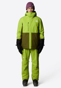 Ljusa gröna vattentäta jacka med bruna paneler, svarta handskar och matchande byxor. Har en hög krage och fickor med dragkedja. Svarta stövlar ingår.