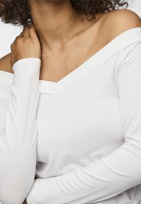 Vit långärmad topp med off-shoulder-design, med en V-formad halsringning och en glatt, mjuk textur. Enkel, minimalistisk stil.