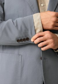 Világosszürke blézer, texturált anyagból, egyetlen zsebbel és négy barna gombbal az ujjnál. Alatta egy bézs csíkos ing található.