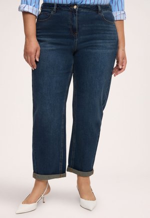 Femme portant un jean bleu foncé retroussé, des escarpins blancs à bout pointu et un chemisier à rayures bleu avec manches roulées.