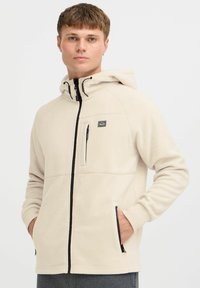 Lys beige fleece hoodie med sort lynlås og detaljer. Har hætte, lynlåslomme på brystet og sidelommer. Glat tekstur.
