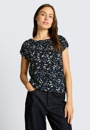 Femme portant un chemisier noir à manches courtes avec des taches blanches abstraites, jean foncé, une main dans la poche, debout devant un fond clair uni.