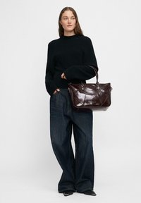 Donna in un maglione nero, jeans a gamba larga scuri e scarpe nere a punta, che tiene una grande borsa in pelle marrone scuro con due manici.