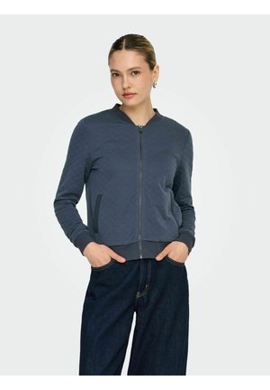 Ženska, ki nosi mornarsko modro prešito jakno z zadrgo, z rebrastim ovratnikom in manšetami, z rokami v žepih, oblečena v temne široke kavbojke.