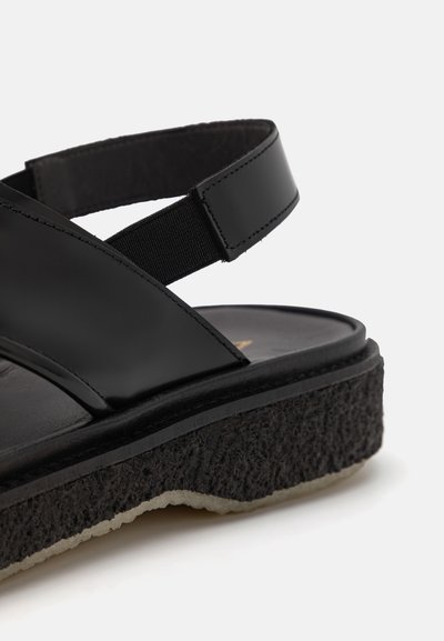 Sandale noire avec des lanières en cuir brillant, un band élastique et une semelle épaisse et texturée de couleur noire avec un bord clair.
