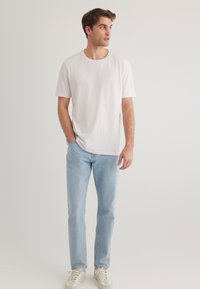White short-sleeve t-shirt with a round neck, paired with light blue jeans and beige sneakers, standing against a plain background.