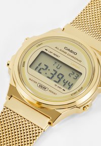 Montre digitale Casio dorée avec un bracelet en maille, affichant l'heure en heures et minutes, avec des boutons pour les réglages de la lumière et de l'alarme.