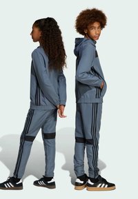Blå idrottsdräkter med svarta ränder, med zip-up huvtröjor och smala byxor. Matchas med svarta och vita sneakers.
