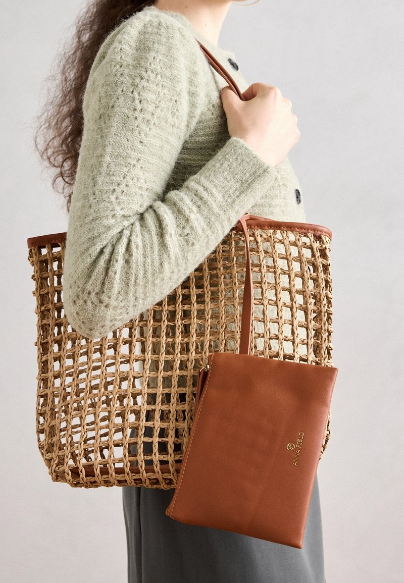 Vävd tote-väska i naturliga fibrer med bruna läderdetaljer, med en fastsydd brun läderpouch. Väskan har en öppen vävdesign.