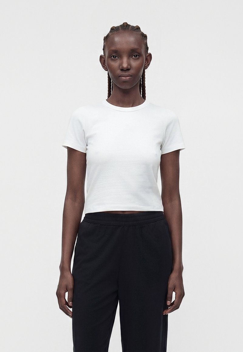 Žena so zopnutými vlasmi, ktorá nosí priliehavý biely krátky crop top s krátkymi rukávmi a voľné čierne nohavice, stojaca pred jednoduchým svetlým pozadím.