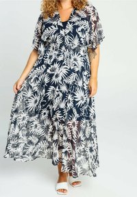 Kvinna som bär en marinblå och vit blommig genomskinlig maxiklänning med korta volangärmar och vita öppna klackar.