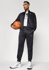 Temno sivi atletski trenirka s jakno na zadrgo in ujemajočimi hlačami, ki imata logotipe Nike. V eni roki drži oranžno košarkaško žogo, obuta v bele superge.