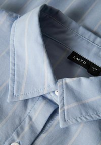 Camicia a righe azzurre chiare con una texture morbida, dotata di colletto a punta, bottoni bianchi e un'etichetta nera con "LMTD" all'interno.
