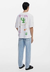 Witte oversized T-shirt met kleurrijke graphics op de achterkant, waaronder een cactus, dierlijke vormen en tekst. Gekocht met lichtblauwe jeans.