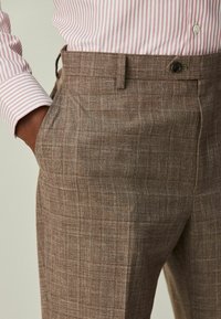 Braune karierte Hose mit subtiler Textur, schmaler Passform und seitlichen Taschen, kombiniert mit einem gestreiften pinkfarbenen Hemd.