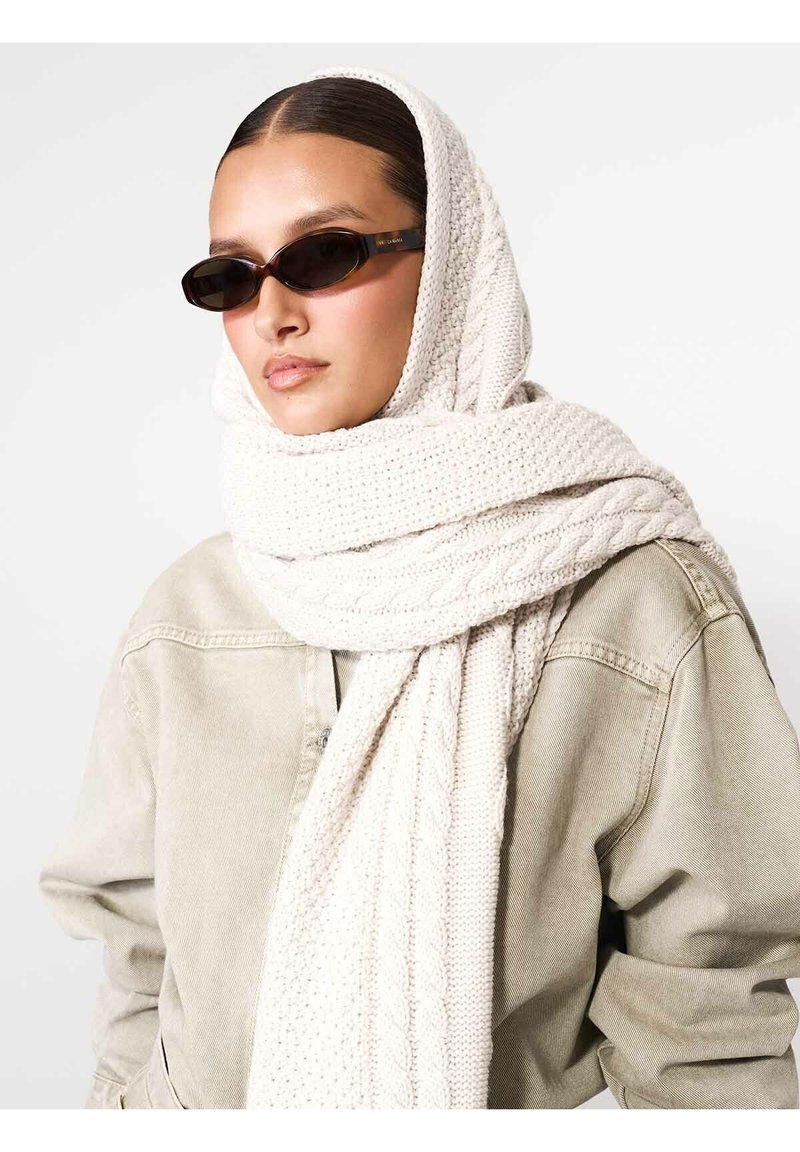 Fular tricotat crem cu textură împletită, suprapus peste o jachetă bej deschis. Modelul poartă ochelari de soare sleek, negri.