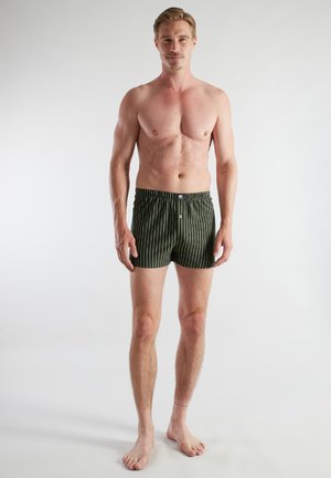 Shirtless man standing barefoot against white background wearing dark green boxer shorts with white vertical stripes.