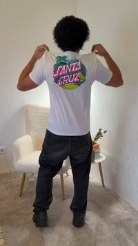 Άτομο με κατσαρά μαλλιά που φοράει λευκό μπλουζάκι "Santa Cruz" και μαύρο παντελόνι, στέκεται μέσα σε κλειστό χώρο κοντά σε μια λευκή πολυθρόνα και ένα μικρό τραπέζι με βιβλία και ένα φυτό.