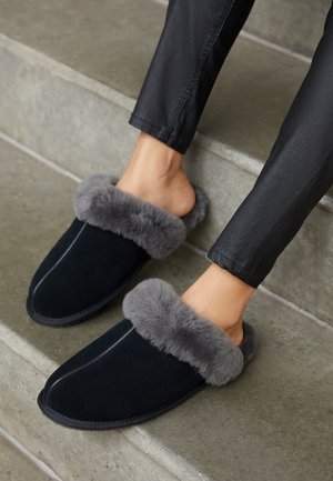 Personne portant des chaussons noirs à enfiler avec une épaisse doublure en fourrure grise, assise sur des marches en béton gris, vêtue d'un pantalon noir.