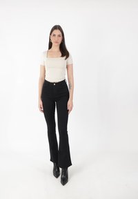 Top beige claro de cuello cuadrado combinado con pantalones vaqueros negros de campana y botas de tobillo negras de punta afilada. Tejido suave y diseño ajustado.