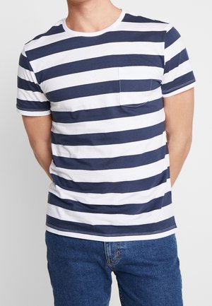 Homme portant un t-shirt à rayures horizontales bleu marine et blanc à manches courtes avec une poche sur la poitrine et un jean bleu, bras derrière le dos.