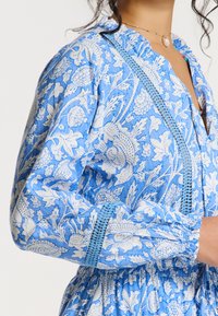 Robe à motifs floraux bleus avec des motifs botaniques blancs, dotée de poignets élastiqués et d'une délicate dentelle le long du décolleté.