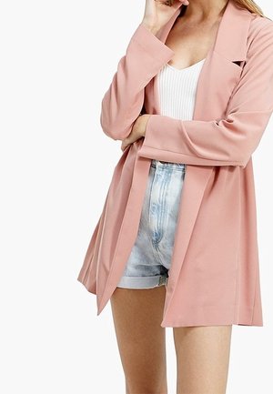 Frau trägt hellrosa Blazer über weißem, geripptem Top und hoch taillierten hellblauen Denim-Shorts, steht mit einem Arm verschränkt und Hand nahe am Gesicht.