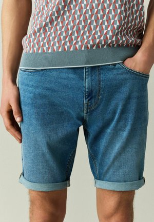 Mann trägt blaue hochgekrempelte Jeans-Shorts und ein gemustertes ärmelloses Top, mit einer Hand in der Tasche und der anderen entspannt an der Seite.