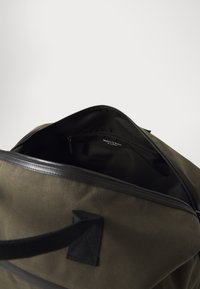 Plátená taška cez rameno v olivovo zelenej farbe s čiernymi koženými detailmi, ktorá má zipsové otváranie a textúrovanú rúčku. Interiér obsahuje malú zipsovú kapsu.