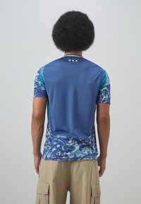 Koszulka sportowa z krótkim rękawem w niebieskiej kolorystyce, z wzorzystą dolną częścią w odcieniach niebieskiego i zielonego oraz żółtymi akcentami przy dekolcie z tyłu.