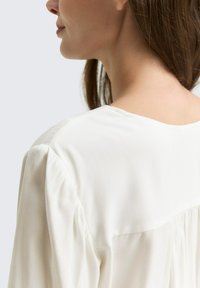 Weißes Blusenshirt aus weichem Stoff, mit lockerem Schnitt, rundem Ausschnitt und puffigen Ärmeln. Glatte Textur, dezente Nahtdetails.