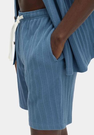 Primer plano de una persona que lleva pantalones cortos de punto acanalado azul con cordón blanco y un cárdigan abierto a juego, con la mano en el bolsillo.