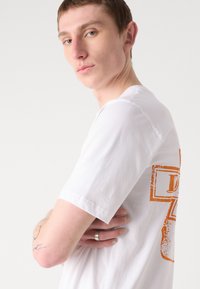 T-shirt bianco in cotone con maniche corte, caratterizzato da un design grafico arancione sul retro. Tessuto morbido, vestibilità comoda, collo a giro.