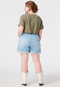 Olijfgroene t-shirt met kantdetails op de schouders, gecombineerd met lichtblauwe denim short met opgerolde mouwen en achterzakken, gedragen met witte cowboylaarzen.