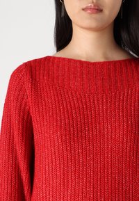 Femme vêtue d'un pull en tricot rouge vif avec un col côtelé et des manches longues, vue des épaules jusqu'au milieu de la poitrine.